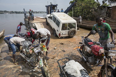 A Toyota 4WD vehicle drives a ferry to cross the Lualaba River. While some moto-taxi drivers are cleaning their bikes in the foreground.