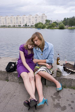 Two women slumped together by the Svislach River. On a summer's evening, people gather on the waterfront where they drink vodka and beer, take paddle boats out for a ride and watch the sunset.