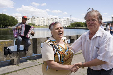 An elderly couple dances to the music of an accordion player. On a summer's evening, people gather on the waterfront where they drink vodka and beer, take paddle boats out for a ride and watch the sun...