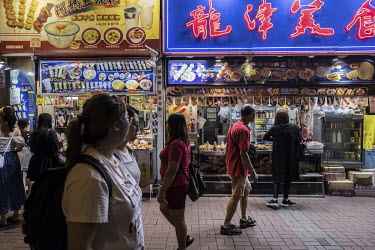 Pedestrians walk past a street food stall with a large neon sign in Mong Kok.
