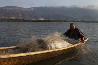 Juan Ixim (70), a Q'eqchi' Mayan fisherman, paddles his boat on Lake Izabal. Behind him, mining activity by CGN-Pronico has left deep scars on Sierra de Santa Cruz.