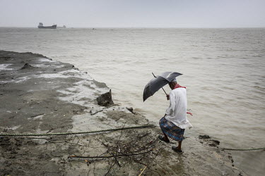 A man walks along the crumbling banks of the Meghna River sheltering from the rain beneath an umbrella. The river has burst its banks during heavy rains devouring land and buildings.
