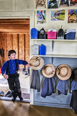 An Amish boy hangs his straw hat on a peg as he entering a classroom at his school.