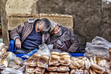 An elderly couple, selling bread from a roadside stall, sleep slumped against each other behind their items.