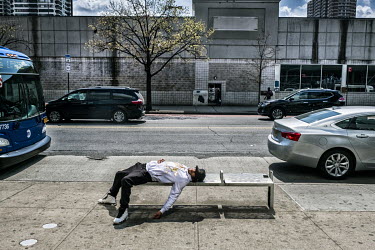 A man, possibly under the influence of drugs or alcohol, sleeps on a bench at a bus stop.