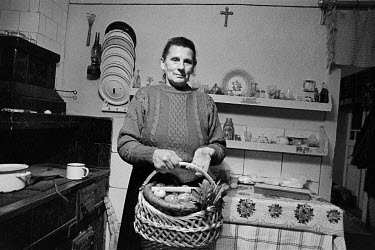 A woman stands in her kitchen holding an Easter basket containing Easter bread (Paska), painted eggs and sausages.