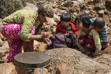 Children, crouched by a woman hunting for rubies and spinels, smile as she shows them what she has found. Children often work with women to help them to find gemstones. As women are not allowed into t...