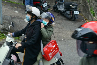 A child, being carried on the back of a motorscooter, adjusts his face mask while travelling through the city.