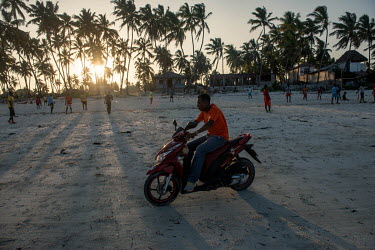 A man rides a motor scooter past a group of men playing football on the beach at Bwejuu.
