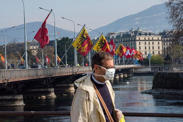 A man wearing a face mask walks near the Pont du Mont Blanc, which is decorated with Swiss and Geneva canton flags, at the point where the lake empties into the Rhone River in the city centre.  Two we...