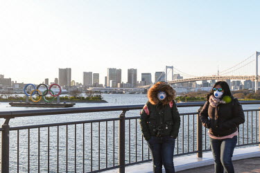 Two women, wearing face masks, stand on the waterside in Tokyo Bay near a barge displaying the Olympic Rings. It has been announced that, due to the coronavirus crisis, the games will be postponed unt...