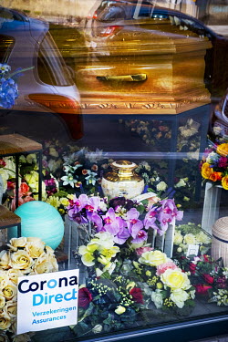 A coffin and plastic flowers are visible in the window of a funeral home. A sticker on the window reads 'Corona Direct Insurance'.