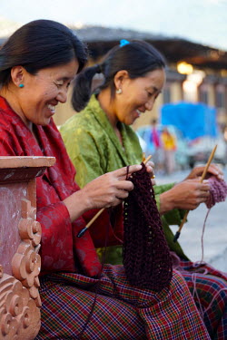 Two women laugh as they sit and knit on a bench in the city centre.