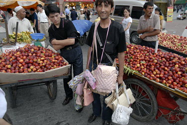 A Uighur man selling handbags beside stalls selling fruits.