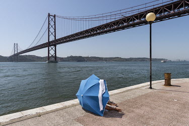 A man shelters from the sun beneath an umbrella near the Ponte 25 de Abril (25th of April Bridge), a suspension bridge connecting Lisbon to the municipality of Almada on the left (south) bank of the T...