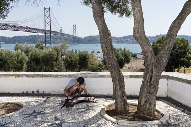A man plays with a dog on a terrace overlooking the Ponte 25 de Abril (25th of April Bridge), a suspension bridge connecting Lisbon to the municipality of Almada on the left (south) bank of the Tagus...