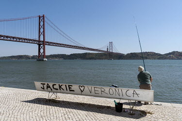 A man fishing near the Ponte 25 de Abril (25th of April Bridge), a suspension bridge connecting Lisbon to the municipality of Almada on the left (south) bank of the Tagus River.