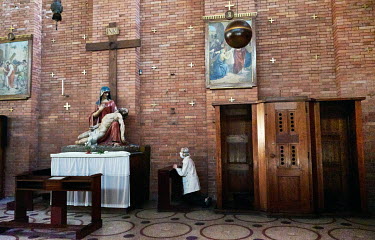 An elderly woman with a face mask praying next to a statue of of Saint Mary holding Jesus with a Cross in backround during Easter Holy Saturday in the Church of the Most Sacred Heart of Our Lord. East...