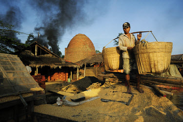 A man carries a yoke with two enormous bamboo baskets filled with rice husks. The husks have been transported by barge to a brick factory where they are used as an alternative to fossil fuels in the b...