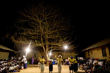 A crowd gathered in the village square to watch a music and dance performance taking place at night, illuminated by solar-powered lights. The villagers have paid to have a solar power system installed...