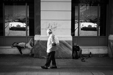 An elderly man walks past two homeless men lying on the ground beside a shop's windows.
