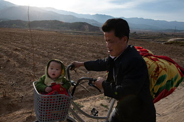 A child sits in a bicycle's front basket while his father wheels it along an unpaved rural road.