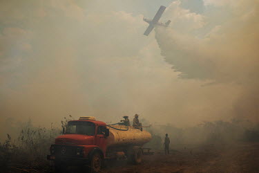 Fire fighters from the Sesc Porto Cercado hotel's private force sit on top of a water truck looking up at an aeroplane as it drops a load of water onto a fire burning near the hotel in the Pantanal.~~...