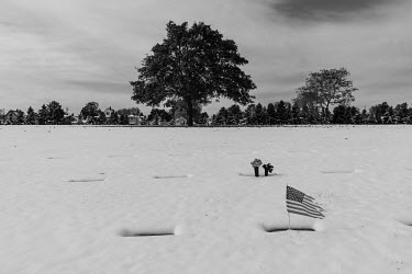 Snow covers the ground at the National Woods Cemetery where a grave is marked by a metal plaque and a mini Stars and Stripes flag.