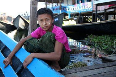 A boy with thanaka sandlewood paste on his face, squats beside a boat moored at the end of a jetty.