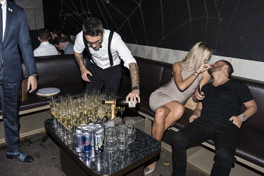 A member of staff pours glasses of champagne into glasses, while beside a couple play about.