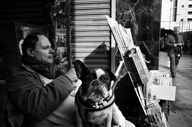 A vendor at a news stand sitting with his dog outside his kiosk.