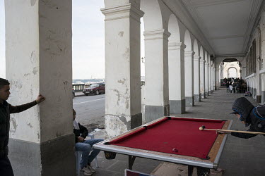 Youths play pool on a table set up within a building's sea front gallery.