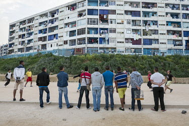 People watch a football match being played on a pitch in front of a residential block.