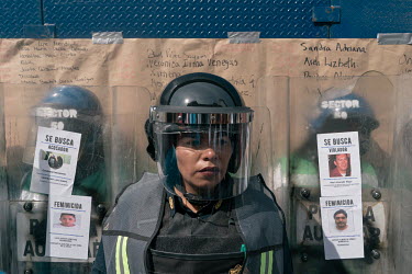 A police officer wearing riot gear stands in front of other police officers holding riot shields on which protestors have attached leaflets displaying the images of men accused of crimes against women...