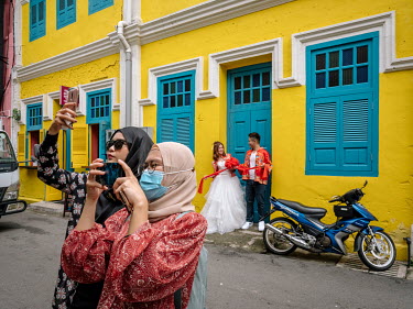 Two veiled women take selfies while behind them a newly wed couple stand in a doorway.