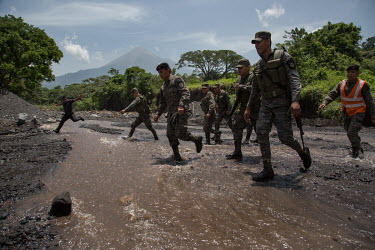 Soldiers cross the Las Casas ravine, one of two fluvial slopes that are crossed to reach the community of 15 de Octubre La Trinidad, as Fuego Volcano erupts in the background. Neither Las Casadas nor...