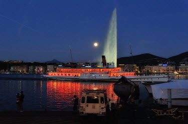 The Belle Epoque ferry boat belonging to the CGN, returning to dock after an evening cruise on the Lake Geneva, passing the illuminated Jet d'eau water feature, and a rising full moon.