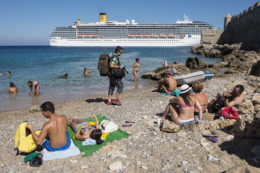 People relax on a beach as behind them a cruise ship arrives in the port of Rhodes.