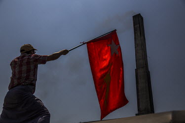 A man waves a National League for Democracy (NLD) flag in salute as smoke rises from a crematorium's chimney during the funeral of Sithu Shein (18) who died when military and police joint security for...