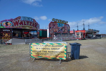A sign explaining COVID-19 restrictions in place at a fun fair (before opening) on waste ground. In the background, the three masts of a sailing vessel at the National Museum of the Royal Navy appear...