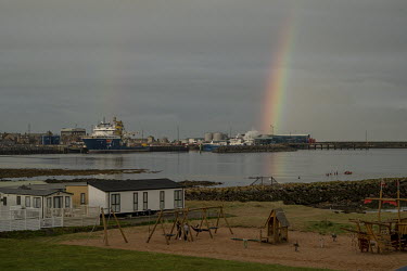 A rainbow rises behind the port at Peterhead, the largest fishing port in the UK, while nearby children ride on swings in a playground.