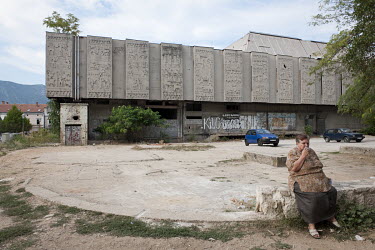 A woman sitting in front of the war-damaged Razvitak Shopping Centre building smoking a cigarette.