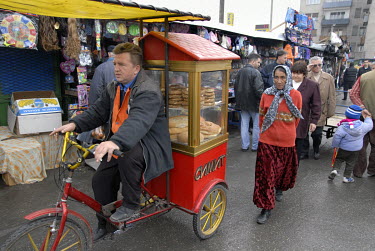 A Roma gypsy woman walks behind a bread seller riding a mobile tricycle stall through the Bit Pazar, a large outdoor market in the predominantly Albanian side of the city.