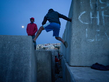 As a full moon rises, a West African migrant looks on as a Moroccan migrant negotiates the concrete sea defences near the port where many young men are camped out trying to evade police after illegall...