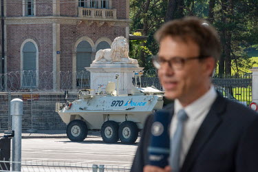 A reporter does a piece to camera near the Joe Biden/Vladimir Putin summit meeting taking place at Villa La Grange. Behind the journalist is an armoured police vehicle.
