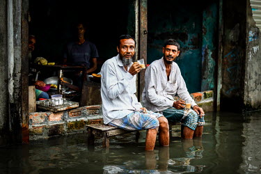 Two men sit drinking tea outside a cafe, their feet in the flood water filling the road.  Water is essential for survival, yet in Bangladesh water also has the power to destroy anything in its path. F...