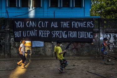 Children play with toy guns in front of a protest banner outside a government office which reads: 'You can cut all the flowers but u can't keep spring from coming.' Soon after soldiers arrived to remo...