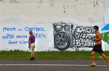 A man stops to read graffiti attacking face masks and vaccines, and proclaiming 'Jesus is Lord', sprayed on a wall along a bicycle path.