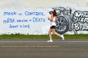 A woman jogging in a face mask passes graffiti attacking face masks and vaccines, and proclaiming 'Jesus is Lord', sprayed on a wall along a bicycle path.