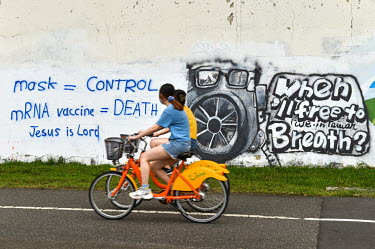 Women ride past graffiti attacking face masks and vaccines, and proclaiming 'Jesus is Lord', sprayed on a wall along a bicycle path.